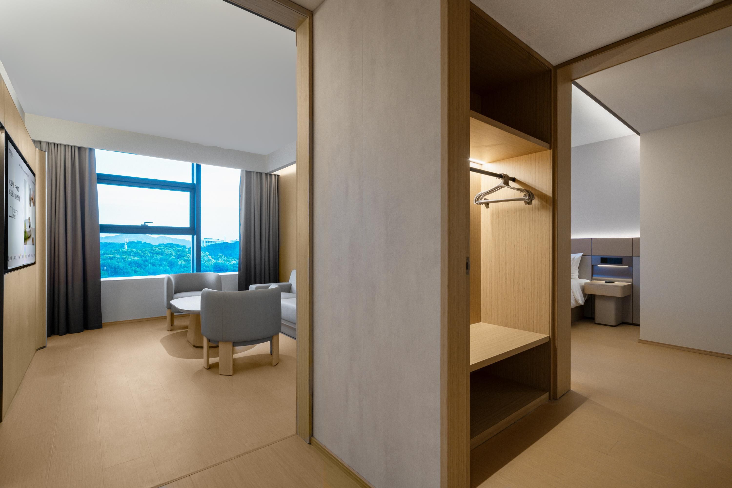 A modern hotel room with a large window, a seating area with two chairs, a wooden wardrobe, and a small bathroom.