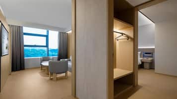 A modern hotel room with a large window, a seating area with two chairs, a wooden wardrobe, and a small bathroom.