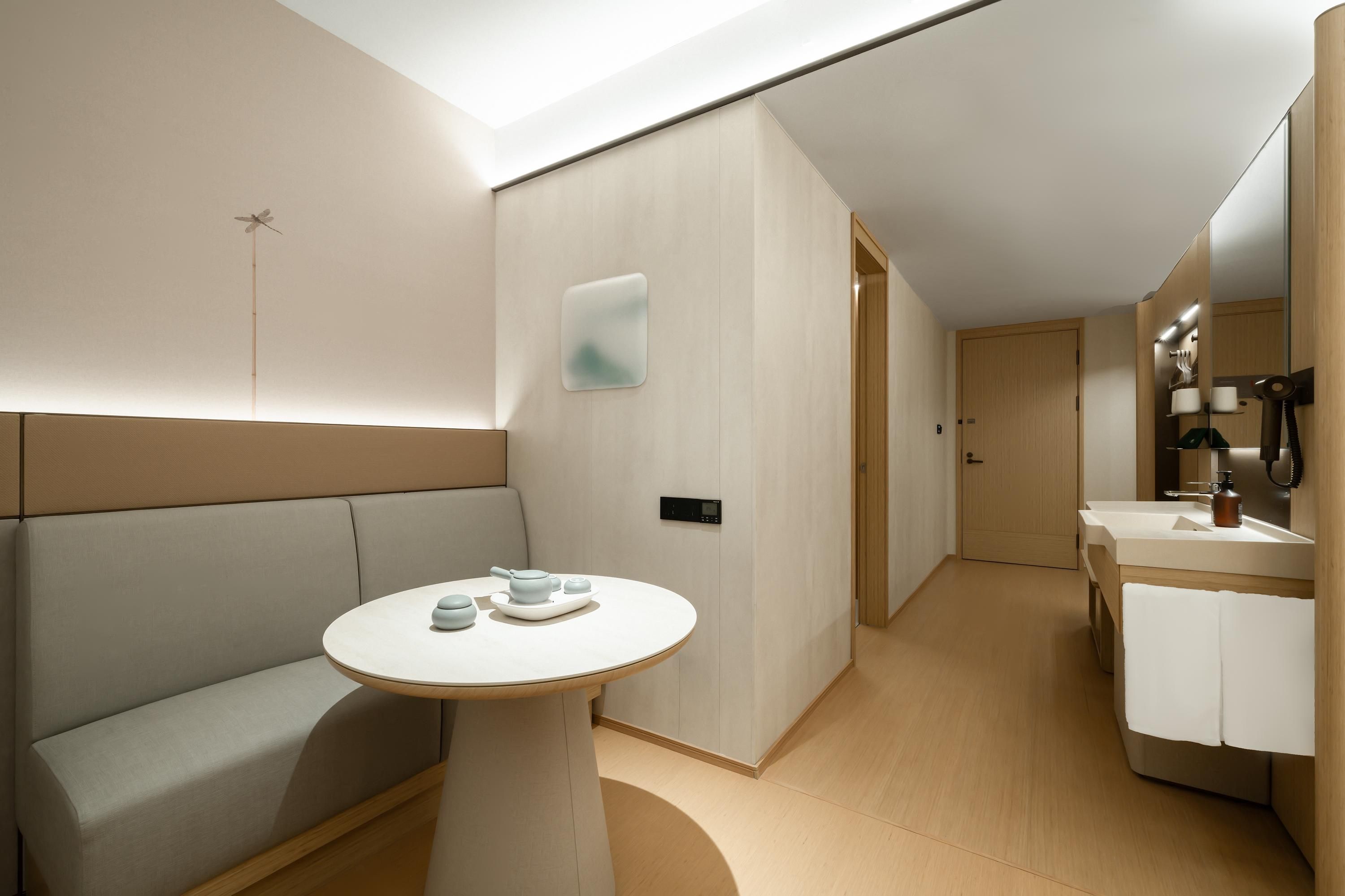 A modern hotel room with a sofa, a round table with teapots, a wall-mounted control panel, and a bathroom with a sink and mirror.