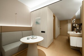 Quarto de hotel moderno com sofá, mesa redonda com bules, painel de controle fixado na parede e banheiro com pia e espelho.