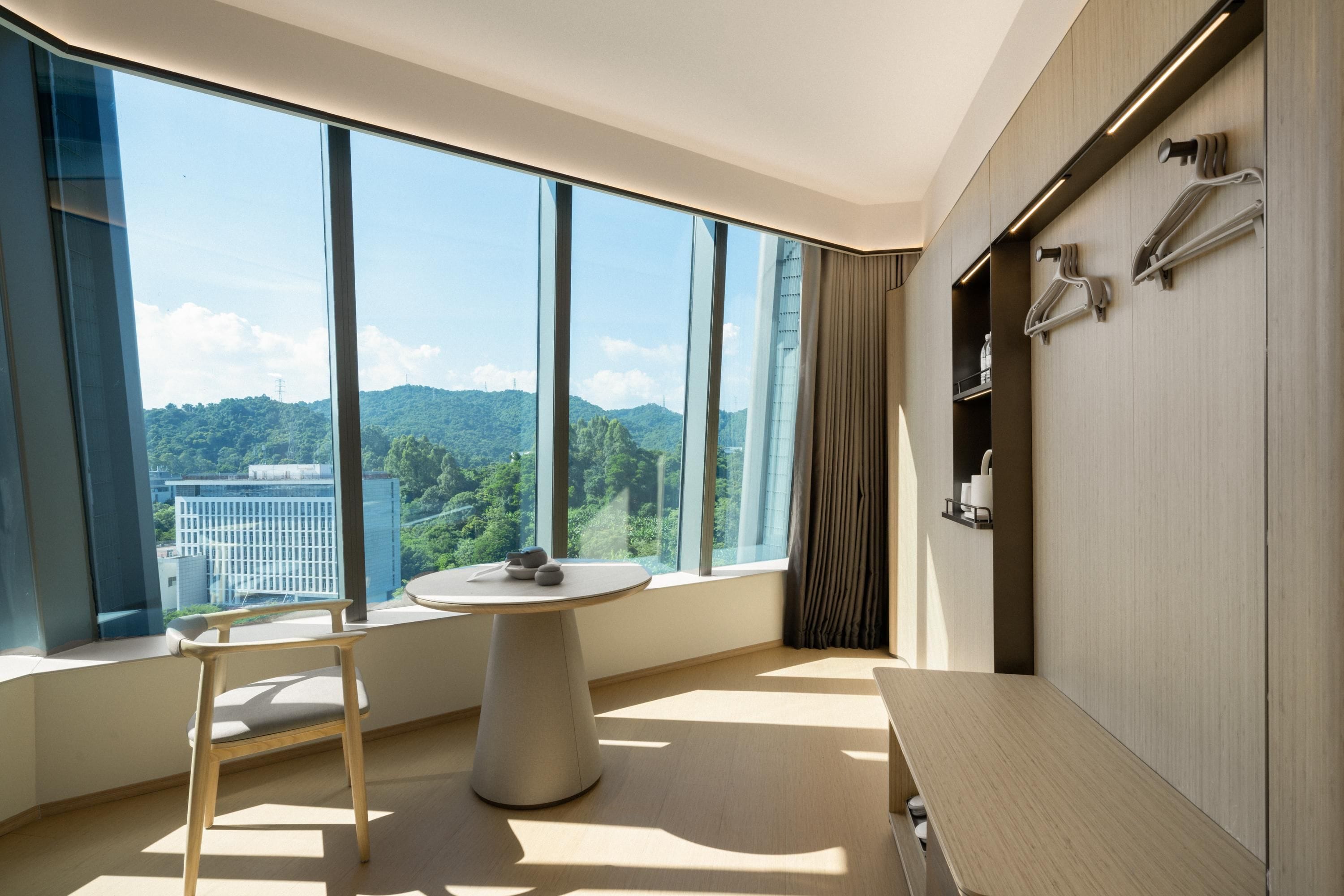 A modern hotel room with large windows, a round table, a chair, a bench, and a wardrobe with hangers.