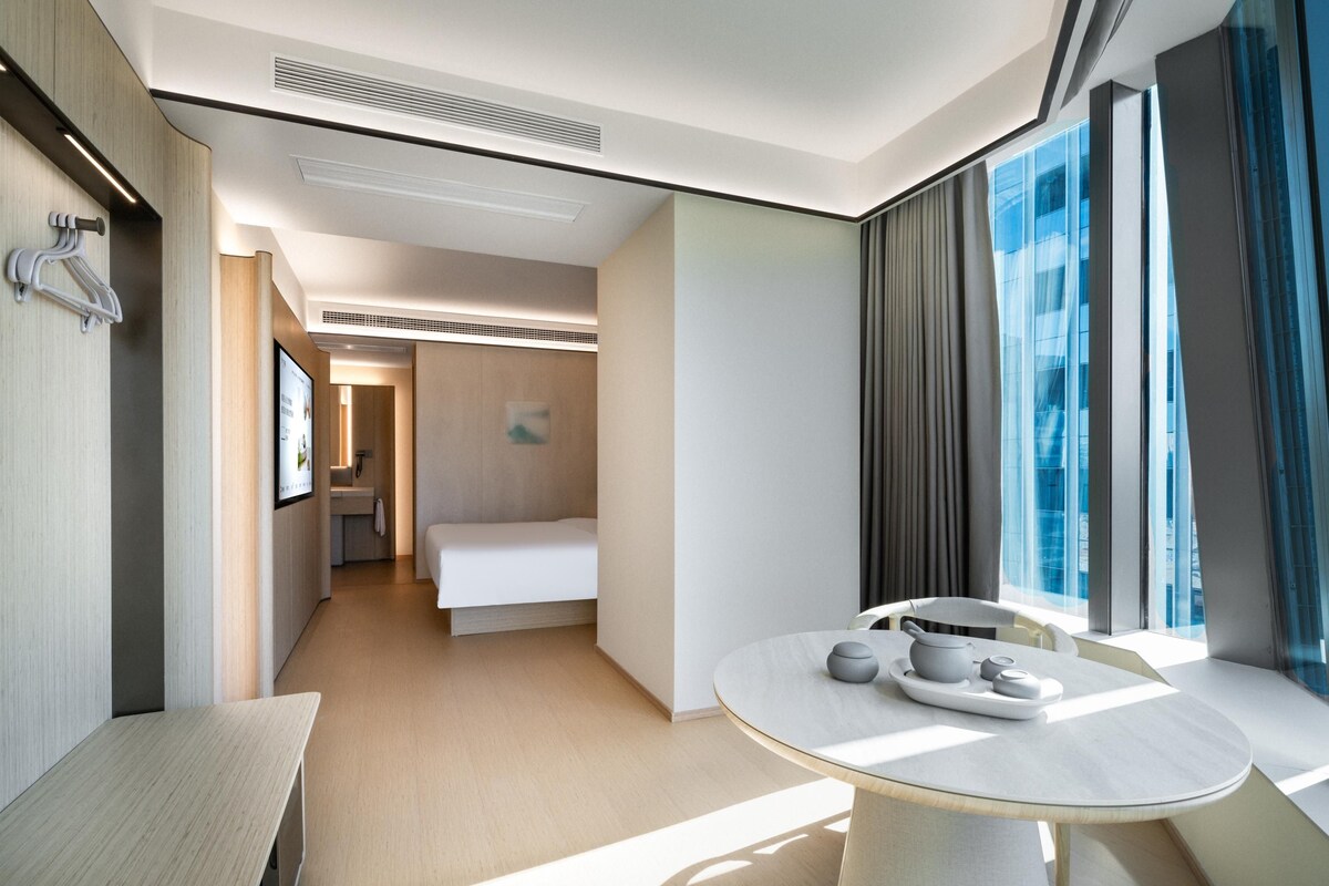 A modern hotel room with a bed, a dining table with a tray, a bench, and a view of the city through large windows.