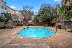 A rectangular pool with a blue interior, surrounded by a pebble-textured deck. There are lounge chairs and a wooden fence in the background.