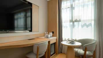 Habitación de hotel con un escritorio de madera, dos sillas, una mesita, televisor y ventana con cortinas.