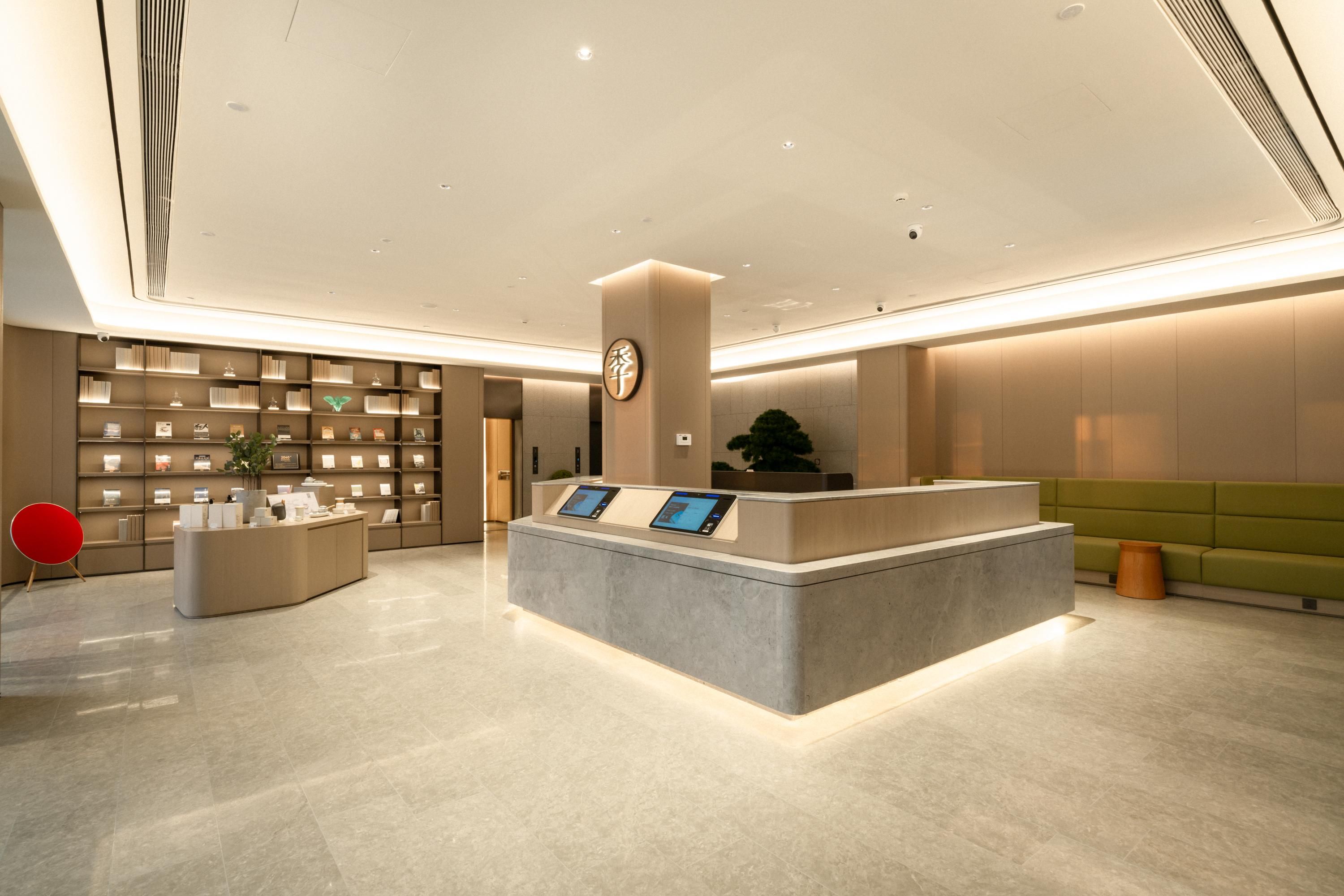 Uma área de recepção de hotel moderna, com um balcão central, um quiosque digital de informações, uma estante de livros e um espaço de estar verde.