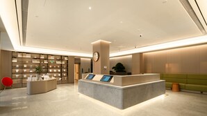 A modern hotel reception area with a central desk, a digital information kiosk, a bookshelf, and a green seating area. - Ji Hotel Guangzhou Huadu Square Metro Station (Guangzhou)