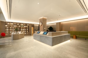 A modern hotel reception area with a central desk, a digital information kiosk, a bookshelf, and a green seating area. - Ji Hotel Guangzhou Huadu Square Metro Station (Guangzhou)