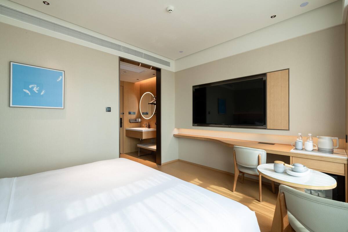 A modern hotel room with a large bed, a desk with a flat-screen TV, a small round table, and a chair.