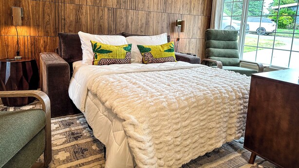 A modern bedroom with a wooden panel wall, a large bed with white bedding, two decorative pillows, a green armchair, and a view of the outdoors through a large window.