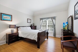 Ein Schlafzimmer mit einem hölzernen Bett, Nachttischen, einem Sessel, einem Fenster mit Vorhängen und Bildern an den Wänden.