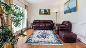 Una sala de estar con un sofá de cuero marrón, un reposapiés a juego, una alfombra floral, un cuadro de un barco en la pared y una planta grande.