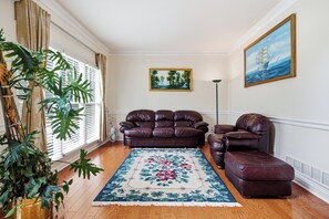 Un salon avec un canapé en cuir marron, un pouf assorti, un tapis à motifs floraux, un tableau représentant un navire accroché au mur et une grande plante.