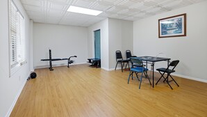 Una habitación con piso de madera, paredes blancas, techo con diseño de cuadrícula, un banco, una mesa con sillas y un cuadro en la pared.