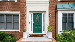 Una casa de ladrillo con una puerta verde, dos plantas en macetas y un pequeño escalón de ladrillo que lleva a la entrada.