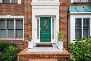 Une maison en briques avec une porte verte, deux plantes en pot et une petite marche en briques menant à l’entrée.