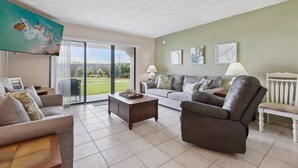 Amplia sala de estar con sofá, sillón y mesa de centro. Hay una puerta corrediza de vidrio que da a un balcón con vistas a zonas verdes.
