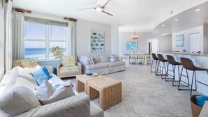 A spacious living area with a large window overlooking the ocean, a modern kitchen with bar stools, and a dining area with a chandelier.