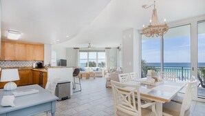 A modern kitchen with wooden cabinets, a dining area with white chairs, and a large balcony with a view of the ocean.