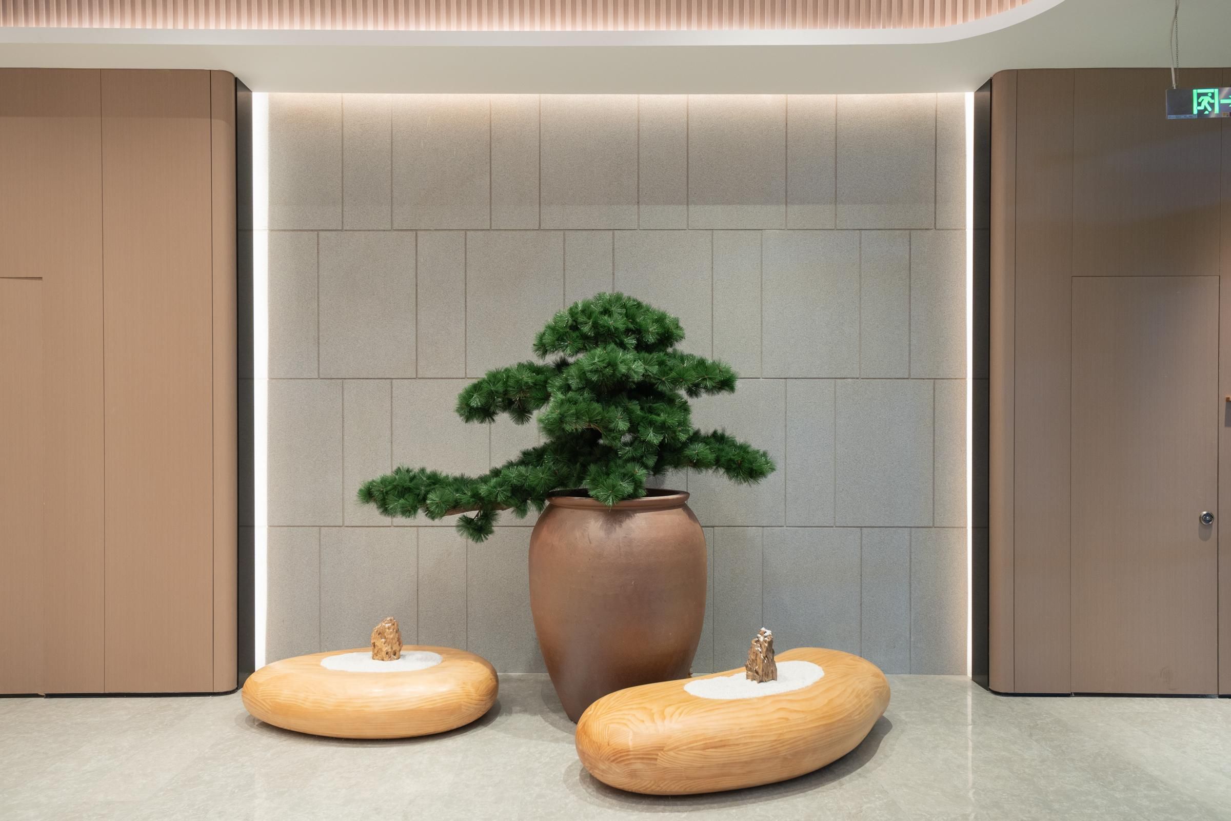 Eine moderne Lobby mit einem großen Kübelbaum, zwei hölzernen Hocker und einer Wand mit eingelassener Beleuchtung.