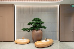 A modern lobby with a large potted tree, two wooden stools, and a wall with a recessed lighting feature.