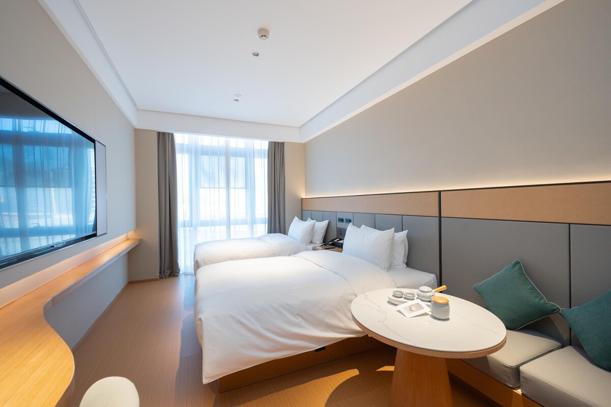 A modern hotel room with a large bed, a sofa, a small table, and a large window.