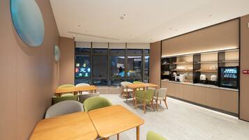 A modern dining area with wooden tables and green chairs, large windows, and a kitchen counter with appliances.