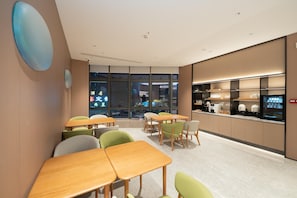 A modern dining area with wooden tables and green chairs, large windows, and a kitchen counter with appliances.