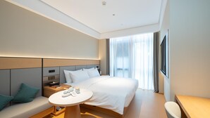 A modern hotel room with a large bed, a bedside table, a small round table, a sofa with cushions, and a large window with curtains. - Ji Hotel Guangzhou Panyu Chimelong Wanbo (Guangzhou)