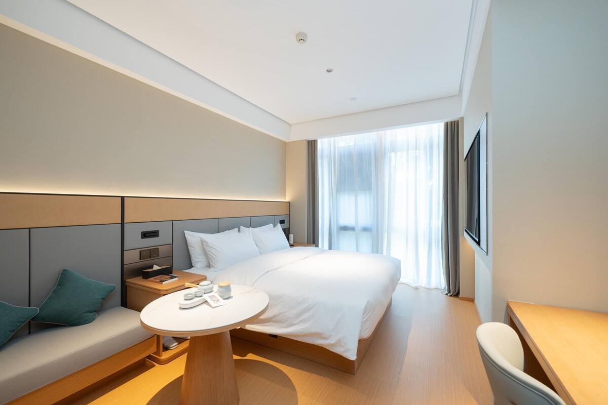 A modern hotel room with a large bed, a bedside table, a small round table, a sofa with cushions, and a large window with curtains.