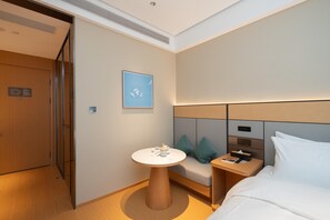 A modern hotel room with a bed, a bedside table with a lamp, a small round table with a teapot, a sofa with cushions, and a wall-mounted TV.