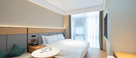 A modern hotel room with a large bed, a bedside table, a small round table, a sofa with cushions, and a large window with curtains.