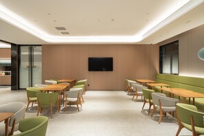A modern dining area with multiple tables and chairs, a flat-screen TV mounted on the wall, and a glass door leading to a kitchen area.