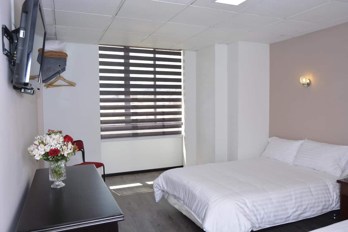 Une chambre d’hôtel avec un lit, un bureau agrémenté d’un vase de fleurs, un téléviseur fixé au mur et une fenêtre avec des stores.