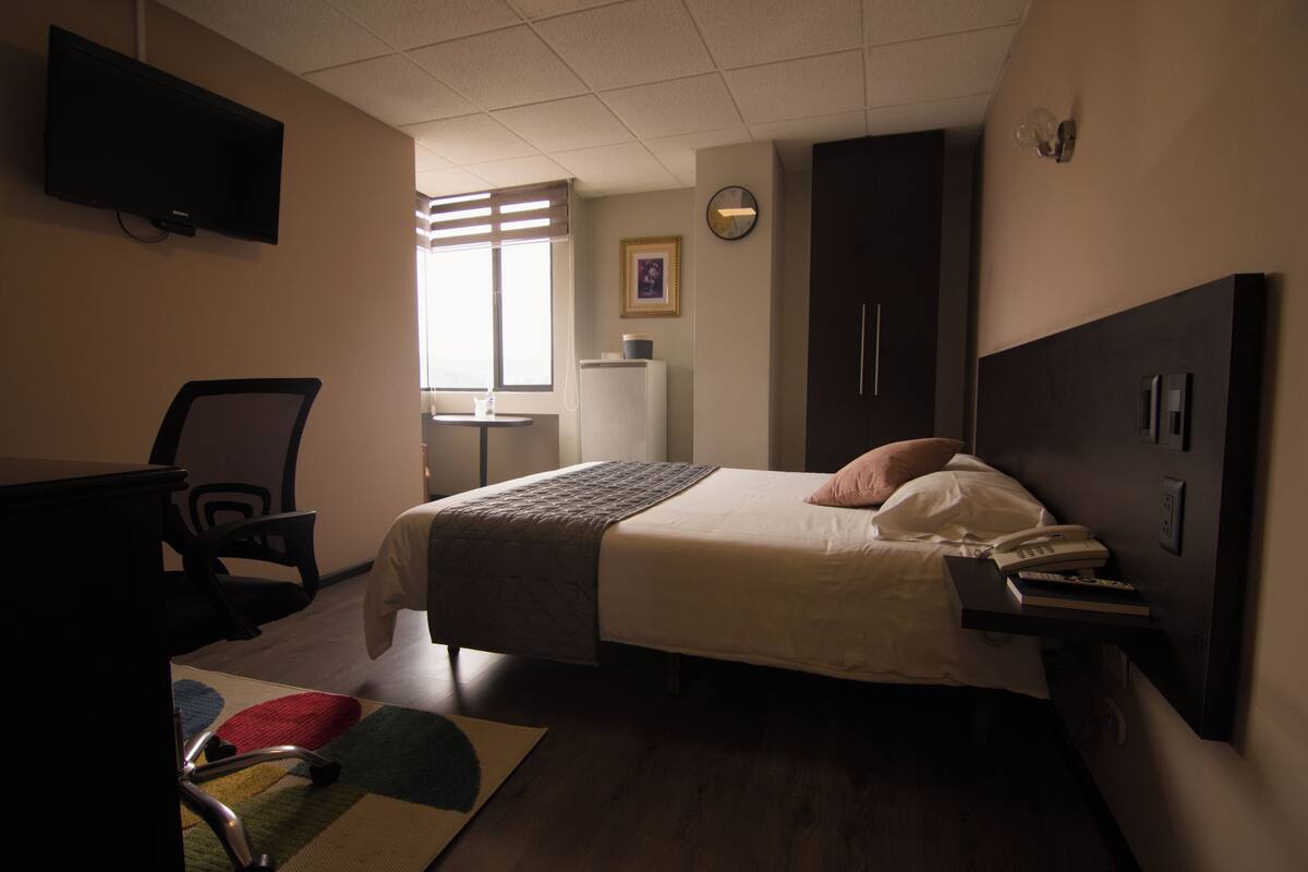 Une chambre d’hôtel équipée d’un lit, d’un bureau, d’une chaise, d’une télévision, d’une horloge et d’une fenêtre avec des stores.
