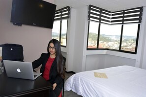A hotel room with a bed, a desk with a laptop, a TV, and a view of a cityscape. - Hotel Italia Cuenca (Cuenca)