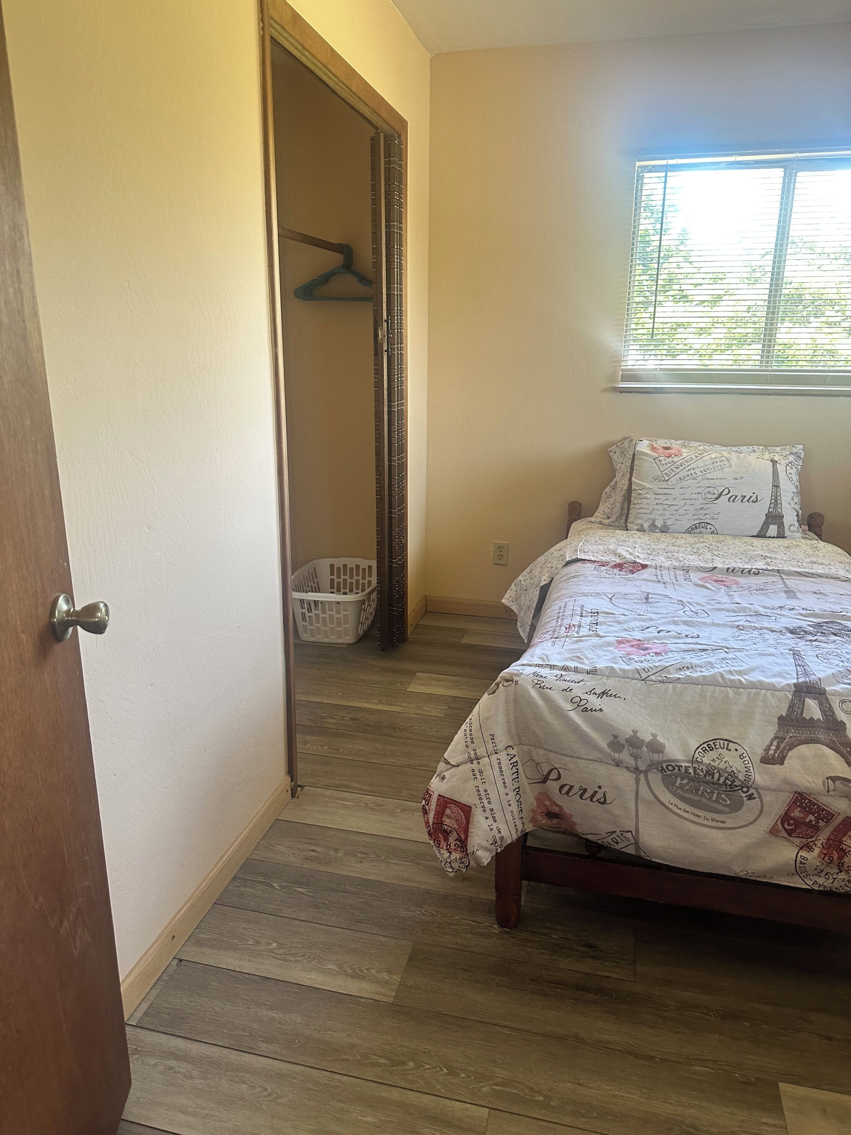 A bedroom with a bed, a window with blinds, a wooden floor, and a door leading to a hallway.