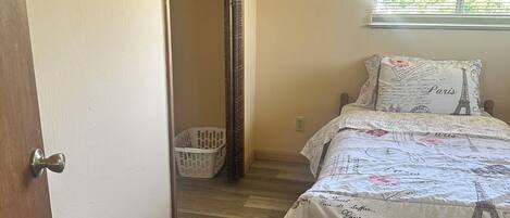 A bedroom with a bed, a window with blinds, a wooden floor, and a door leading to a hallway.