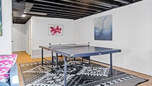 A ping pong table in a room with white walls and a black ceiling. There are two paintings on the walls, one with a pink flower and the other with blue abstract patterns. The floor has patterned rugs. - Completely Renovated Home in St. Charles, Missouri (Saint Charles)