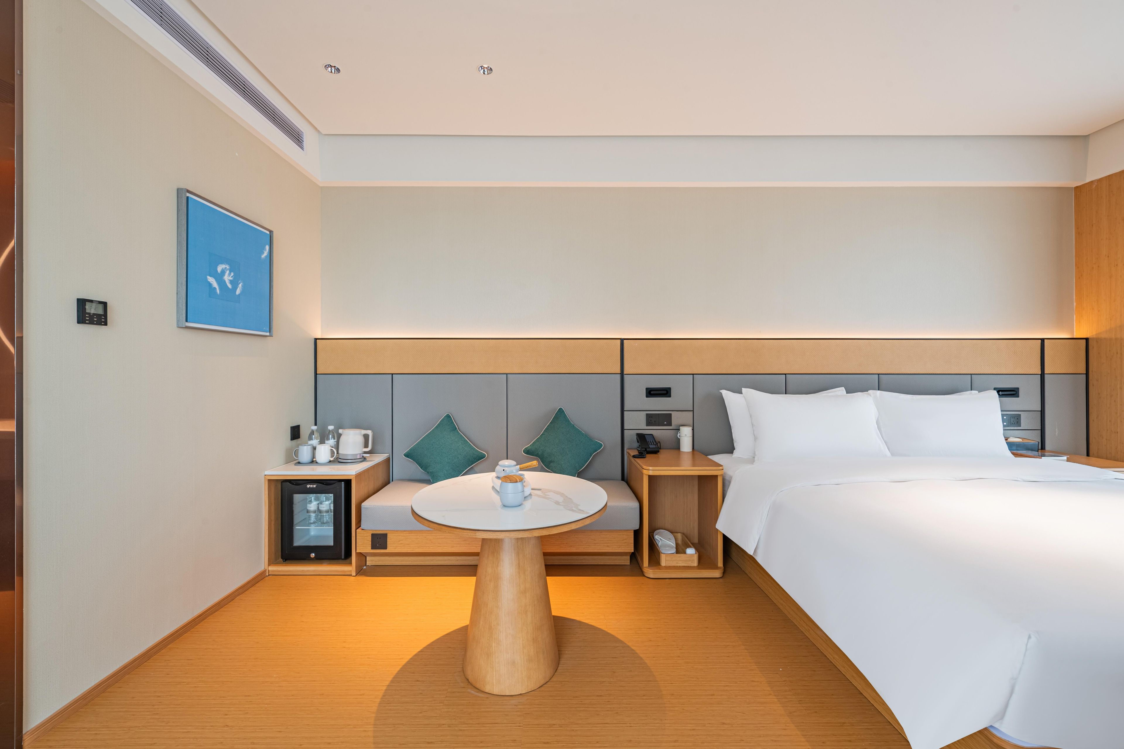 A modern hotel room with a bed, a round wooden table, a small fridge, a bedside table with a lamp, and a wall-mounted TV.