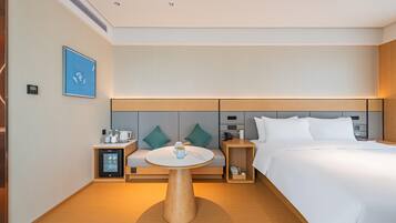 A modern hotel room with a bed, a round wooden table, a small fridge, a bedside table with a lamp, and a wall-mounted TV.