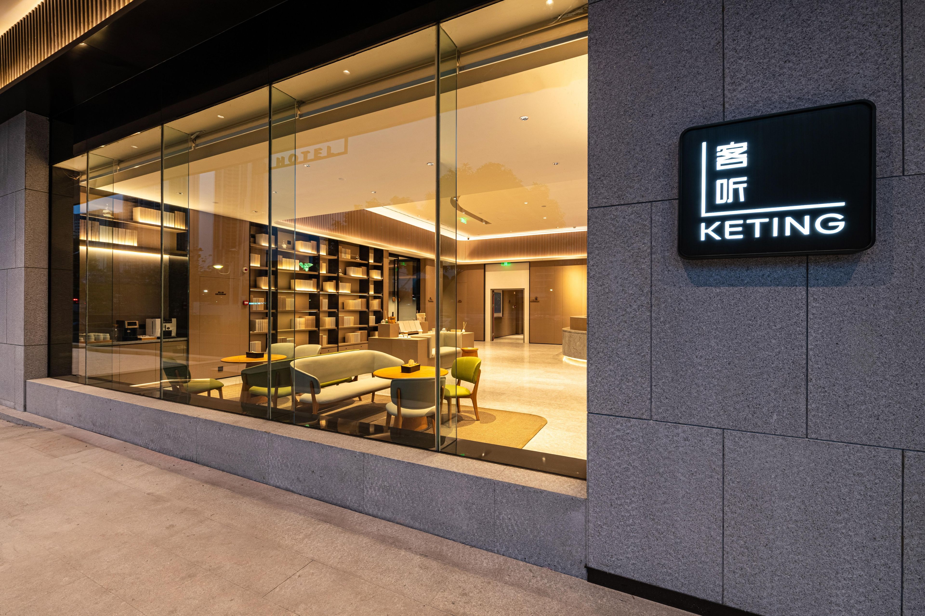 Interior moderno de hotel com fachada de vidro, uma placa indicando 'KETING' e área de estar com cadeiras e mesas.