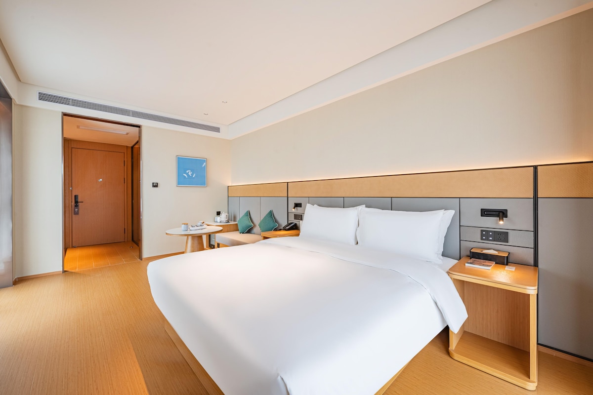 A modern hotel room with a large bed, bedside tables, a nightstand, and a door leading to another room.