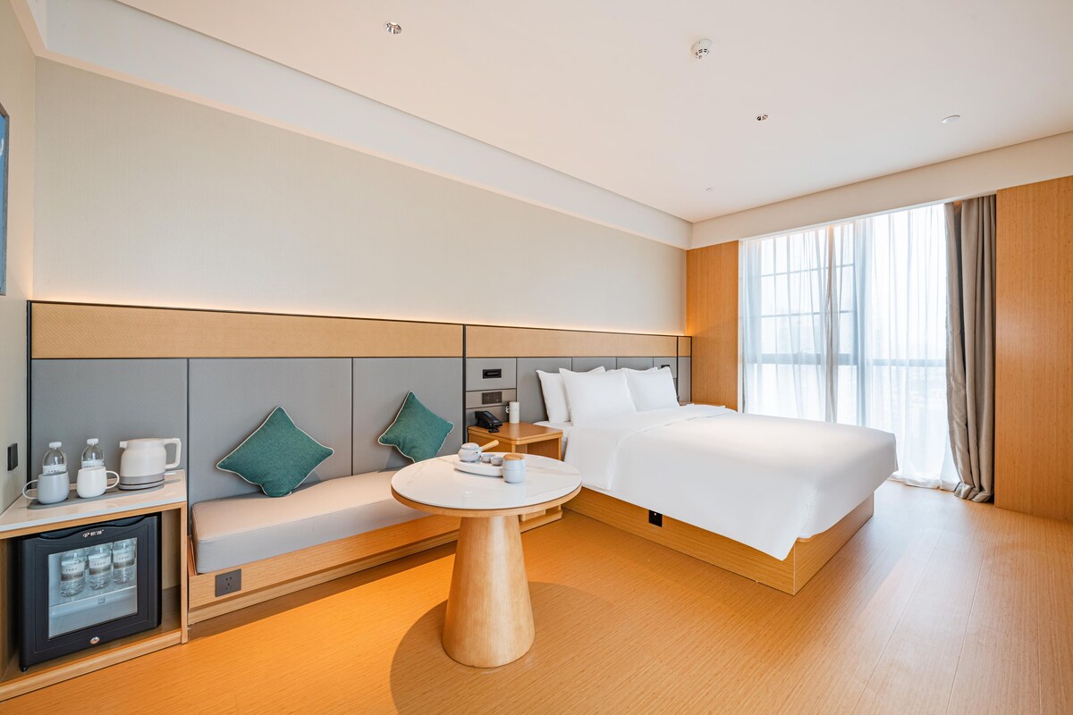 A modern hotel room with a large bed, a seating area with a table, and a minibar.
