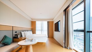 Habitación de hotel moderna con una cama grande, una mesita de noche, un sofá, un televisor y una ventana con vista a la ciudad.