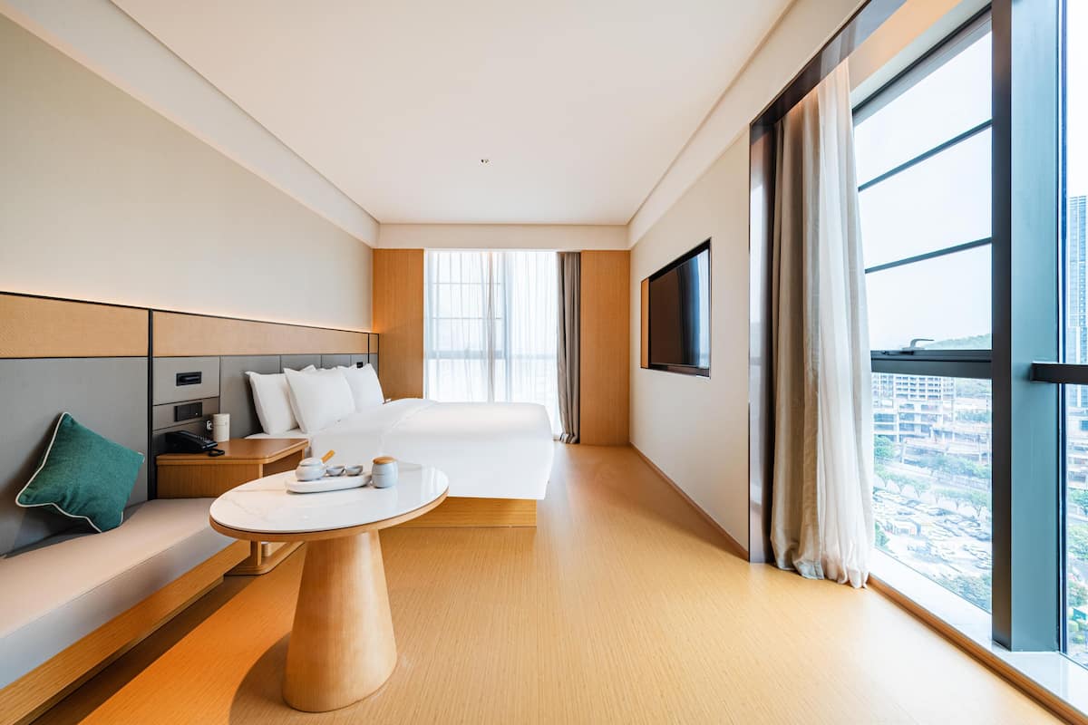 Ein modernes Hotelzimmer mit einem großen Bett, einem Nachttisch, einer Couch, einem Fernseher und einem Fenster mit Blick auf die Stadt.
