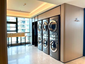 A modern laundry room with four front-loading washing machines, a small table with baskets, and a large window providing a view of the building's exterior. - Ji Hotel Guangzhou Panyu Shiqiao Metro Station (Guangzhou)