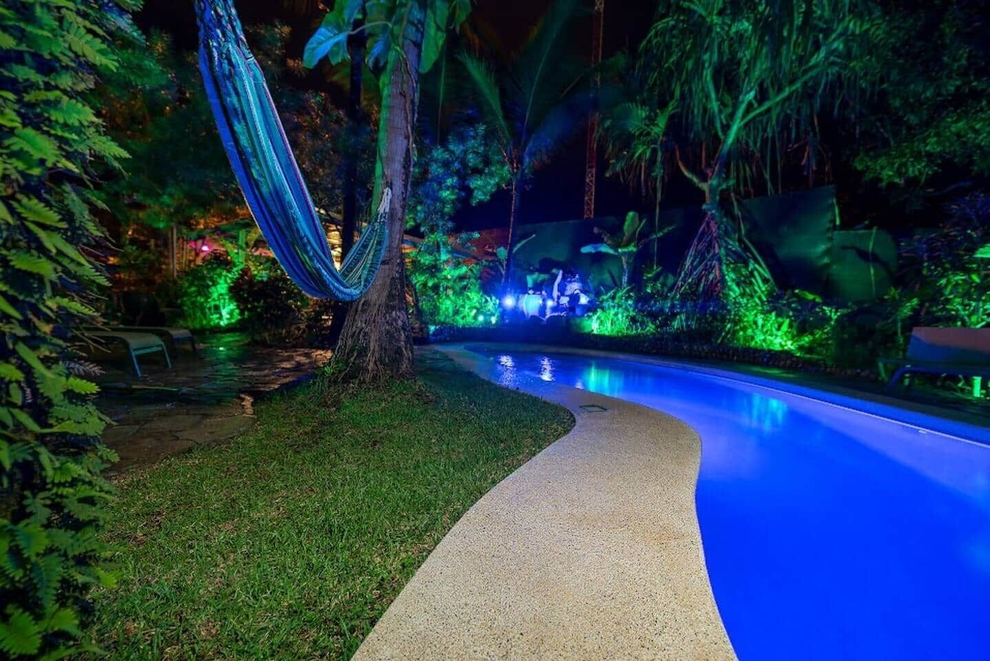 A nighttime pool area with a hammock, surrounded by lush greenery and illuminated by blue and green lights.