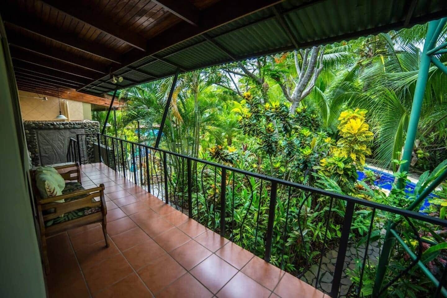Un balcon doté d’un sol carrelé, d’une chaise en bois et offrant une vue sur une végétation luxuriante et une piscine.