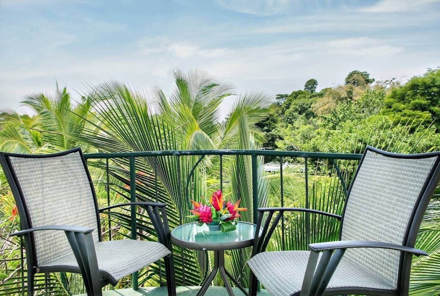 A balcony with two chairs and a table, surrounded by lush greenery and a clear sky.