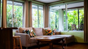 Una sala de estar con un sofá, una mesa de centro de madera y vistas a abundante vegetación a través de amplios ventanales.